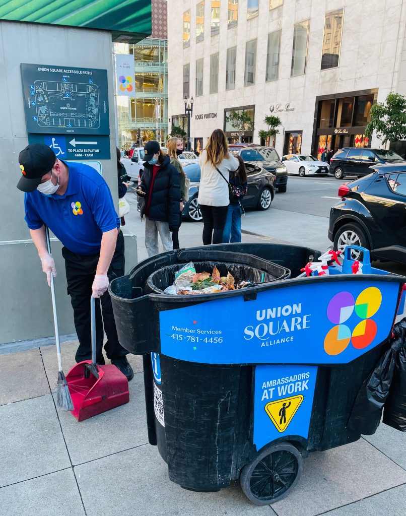 A Block by Block employee in a blue uniform sweeps the sidewalk in Union Square.