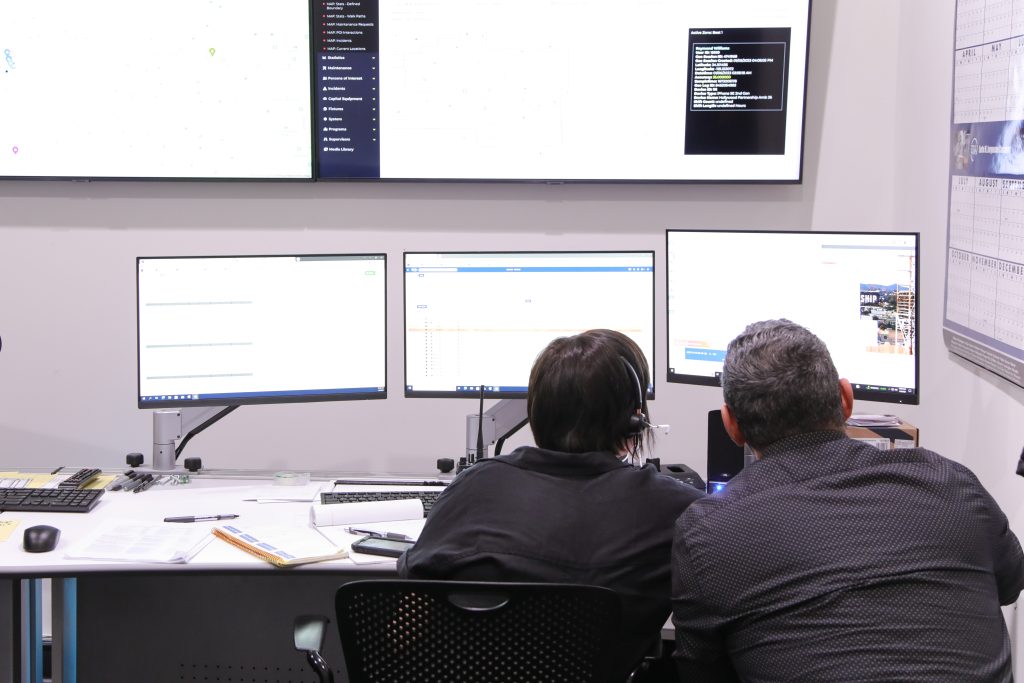 Two people in black look at three computer screens.